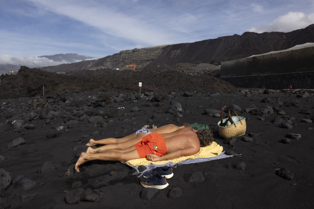 La Bombilla. A German couple who have been living on the island for over 20 years sunbathe on the edge of the beach infront of the new lava flow which has overtaken the once long beach and formed a new peninsula.  It is forbidden to walk on the lava field due to risk of collapse and gas emissions.  

La Palma. An island of the Canaries. Two years after the Cumbre Vieja volcanic eruption in late 2021.  Photography taken on January 2024.